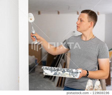 Young man painting wall with paint roller in apartment during repair works Young man painting wall with paint roller in apartment during repair works 119608196