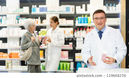Portrait of friendly man druggist in lab coat standing in drugstore with drug package Portrait of friendly man druggist in lab coat standing in drugstore with drug package 119608250