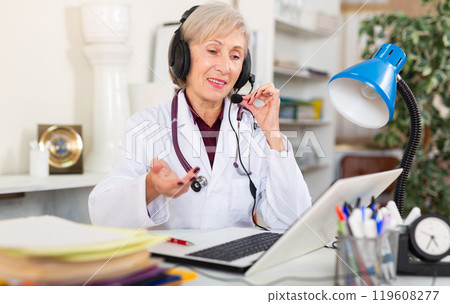 Smiling aged female doctor in headset conducting online consultation Smiling aged female doctor in headset conducting online consultation 119608277