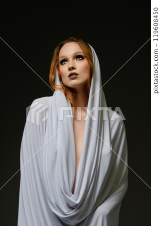 Ethereal portrait of a woman in a flowing white drape against a dark backdrop 119608450