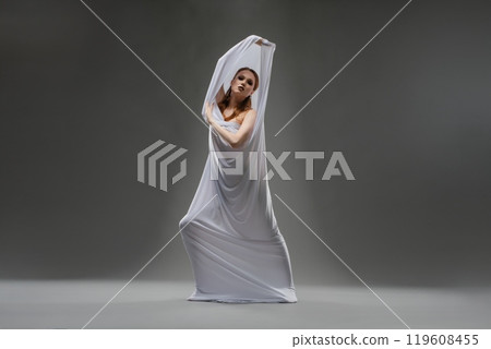 A dancer wearing a flowing white fabric performs a graceful pose indoors 119608455
