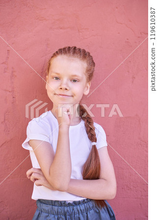 Confident young girl with red hair posing against a pink wall in casual outfit 119610387