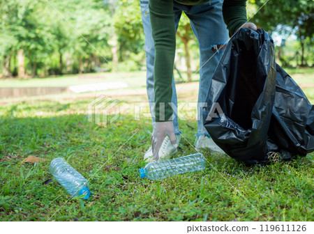 Woman of dedicated volunteers are participating in an environmental cleaning activity in a park. Woman wearing gloves and hold large black bags. Woman collecting trash and plastic bottles in a park. Woman of dedicated volunteers are participating in an environmental cleaning activity in a park. Woman wearing gloves and hold large black bags. Woman collecting trash and plastic bottles in a park. 119611126