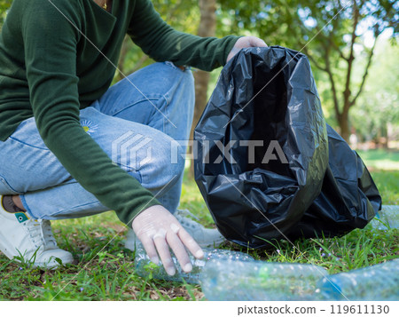 Woman of dedicated volunteers are participating in an environmental cleaning activity in a park. Woman wearing gloves and hold large black bags. Woman collecting trash and plastic bottles in a park. Woman of dedicated volunteers are participating in an environmental cleaning activity in a park. Woman wearing gloves and hold large black bags. Woman collecting trash and plastic bottles in a park. 119611130
