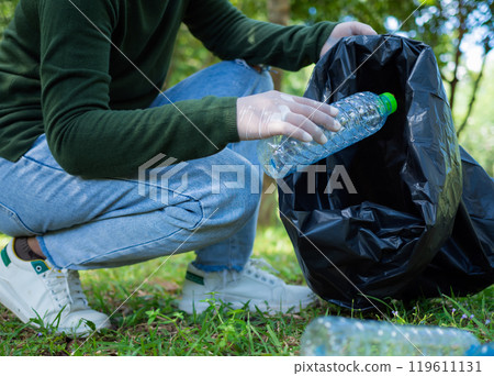 Woman of dedicated volunteers are participating in an environmental cleaning activity in a park. Woman wearing gloves and hold large black bags. Woman collecting trash and plastic bottles in a park. Woman of dedicated volunteers are participating in an environmental cleaning activity in a park. Woman wearing gloves and hold large black bags. Woman collecting trash and plastic bottles in a park. 119611131