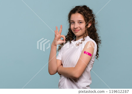 Girl making "OK" gesture with pink bandage on vaccinated arm. 119612287