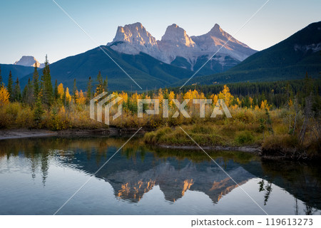 Beautiful scenery of the Canadian Rockies at dawn in autumn. Mountain, forest in yellow and green color reflected on water. Canmore, Alberta, Canada. The Three Sisters trio of peaks. Beautiful scenery of the Canadian Rockies at dawn in autumn. Mountain, forest in yellow and green color reflected on water. Canmore, Alberta, Canada. The Three Sisters trio of peaks. 119613273
