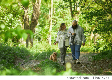 Couple casually walking their dog through vibrant green forest, enjoying nature. Both individuals smile while holding hands and leash Couple casually walking their dog through vibrant green forest, enjoying nature. Both individuals smile while holding hands and leash 119615764