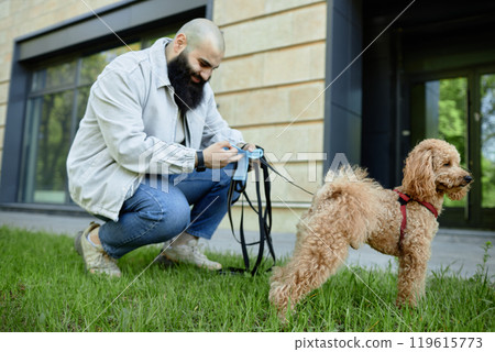Bearded man kneeling on grass while tending to small poodle's collar in front of modern building with large windows Bearded man kneeling on grass while tending to small poodle's collar in front of modern building with large windows 119615773