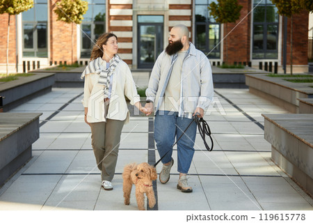 Couple walking hand in hand outdoors with their dog on leash, smiling and looking at each other with happiness, dressed in casual clothes on sunny day with modern buildings in background 119615778