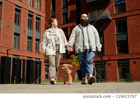 Couple holding hands while walking their dog in modern residential neighborhood with red brick buildings surrounding them showcasing contemporary architecture Couple holding hands while walking their dog in modern residential neighborhood with red brick buildings surrounding them showcasing contemporary architecture 119615787