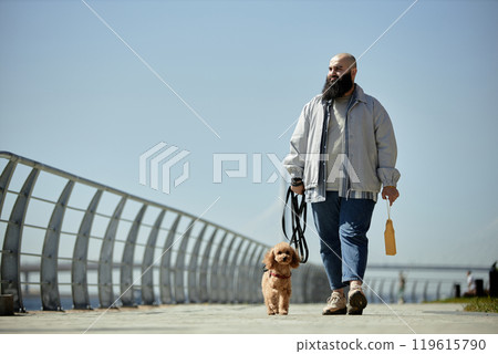 Man walking small brown dog along a path bordered by curved railings against clear blue sky holding bag in hand 119615790