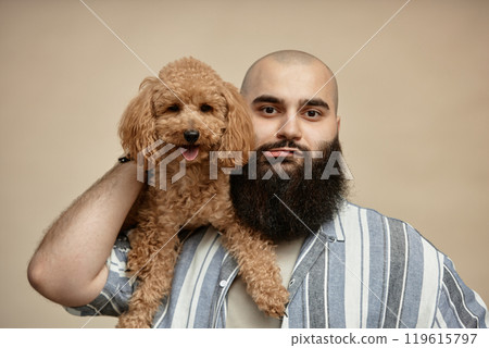 Portrait of bearded man holding curly-haired dog against solid background, wearing striped shirt, creating a friendly and inviting atmosphere Portrait of bearded man holding curly-haired dog against solid background, wearing striped shirt, creating a friendly and inviting atmosphere 119615797