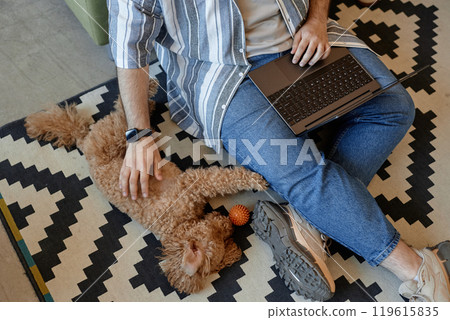 Casual setting showing person using laptop while sitting on floor with fluffy dog lying beside. Person wearing striped shirt and blue jeans, and seated on patterned rug 119615835
