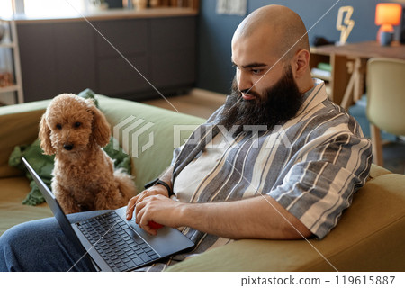 Bearded man sitting on couch typing on laptop while small dog intently watching beside, creating a cozy and focused atmosphere in a modern living room 119615887