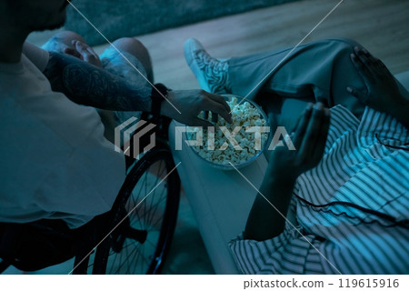 Two friends sharing popcorn during an evening indoor gathering, one sitting in wheelchair and the other reaching into bowl from couch, engaging in relaxed conversation 119615916