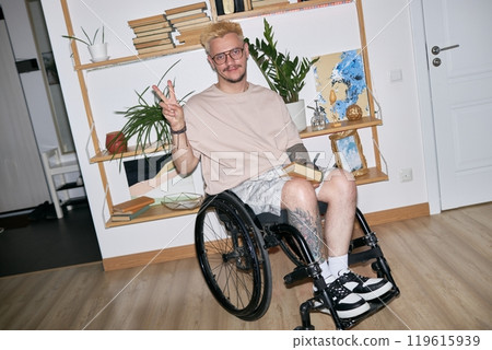 Man sitting in wheelchair and smiling while holding book and flashing peace sign in well-decorated room with plants 119615939