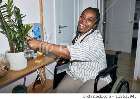Woman sitting in wheelchair, with joyful expression, watering potted plant. Contemporary home interior with wooden shelves, artwork, and door visible Woman sitting in wheelchair, with joyful expression, watering potted plant. Contemporary home interior with wooden shelves, artwork, and door visible 119615943