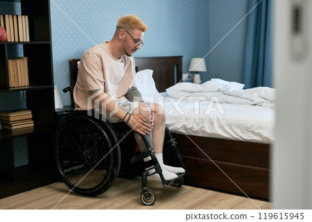 Man sitting in wheelchair adjusting prosthetic leg beside unmade bed in cozy bedroom. Bookshelf filled with various books visible in background Man sitting in wheelchair adjusting prosthetic leg beside unmade bed in cozy bedroom. Bookshelf filled with various books visible in background 119615945