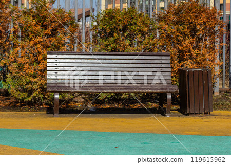 Empty wooden bench on a sunny day. Cozy wooden bench on a colorful sidewalk in a quiet residential area surrounded by trees and bushes, offering a peaceful rest in the city. Empty wooden bench on a sunny day. Cozy wooden bench on a colorful sidewalk in a quiet residential area surrounded by trees and bushes, offering a peaceful rest in the city. 119615962