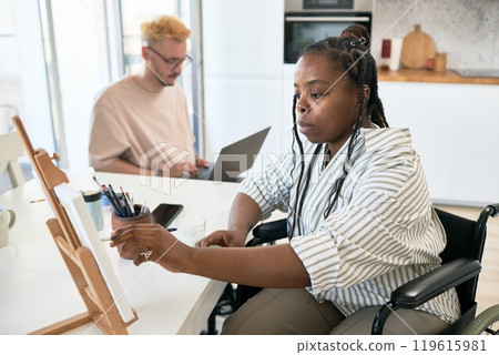 Two individuals engaging in creative work in home office setting, one painting on canvas while other works on laptop in background 119615981