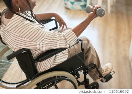 Person in wheelchair exercising with dumbbell in cozy interior. Hand has relaxed grip on weight while focused on activity, creating positive, motivated atmosphere 119616092