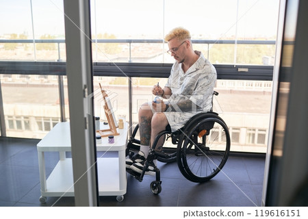 Man, using wheelchair, is engrossed in painting at table near window in bright room. His art supplies are neatly arranged on nearby table, and he concentrates on his work, brush in hand Man, using wheelchair, is engrossed in painting at table near window in bright room. His art supplies are neatly arranged on nearby table, and he concentrates on his work, brush in hand 119616151