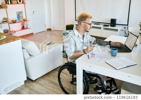 Man sitting in his wheelchair, working on laptop at home office desk, surrounded by papers, notes, and coffee cup. Modern, clean living space visible with shelves and decor 119616193