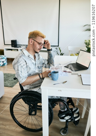 Man in wheelchair at home office desk with documents and laptop, focusing on work during daytime. Coffee mug, pen, and notebook on desk, creating productive workspace Man in wheelchair at home office desk with documents and laptop, focusing on work during daytime. Coffee mug, pen, and notebook on desk, creating productive workspace 119616207