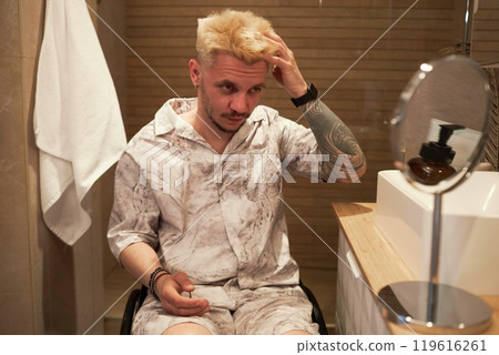 Man with tattoos on both arms, sitting in bathroom, examining hairstyle in mirror, wearing patterned pajamas, displaying focused demeanor during grooming routine Man with tattoos on both arms, sitting in bathroom, examining hairstyle in mirror, wearing patterned pajamas, displaying focused demeanor during grooming routine 119616261