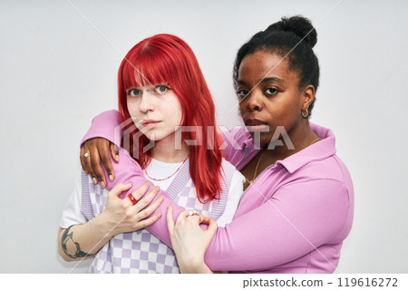 Portrait of red-haired Caucasian woman and African American woman standing close with arms around each other, expressing solidarity and friendship, both wearing purple and white attire Portrait of red-haired Caucasian woman and African American woman standing close with arms around each other, expressing solidarity and friendship, both wearing purple and white attire 119616272