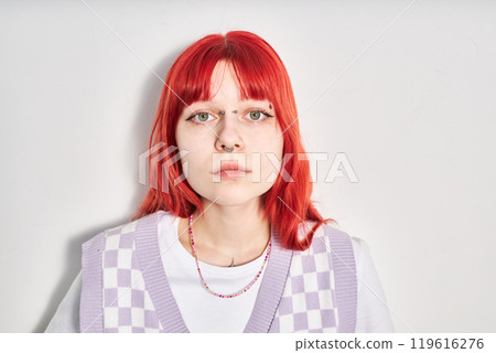 Portrait of young woman with bright red hair and piercing, wearing purple checkered vest and white shirt, standing against white wall, looking straight at camera 119616276