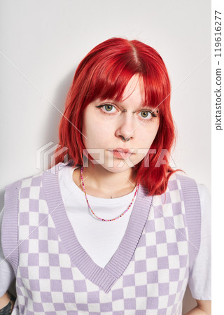 Portrait of young woman with vibrant red hair wearing a checked purple vest over white shirt and bead necklace, straight at camera with neutral expression 119616277