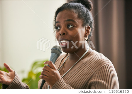 Young African American woman giving speech while holding microphone and gesturing with hand Demonstrating expression of engagement and confidence 119616350