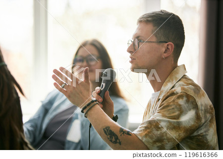 Young man with glasses speaking into microphone during conference event with blurred attendees in background. Wearing casual attire and showcasing engaged facial expression 119616366