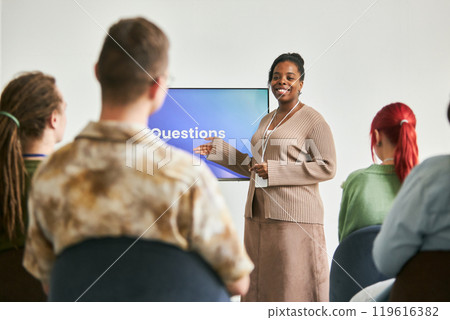 Smiling African American woman presenting to diverse group with large display screen in background wearing beige sweater and holding pen while answering questions 119616382