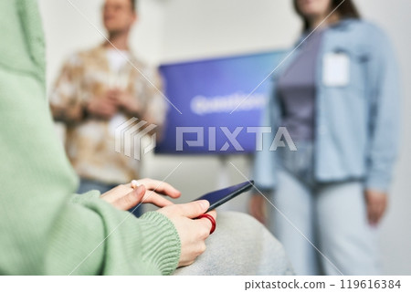 Close-up of person holding smartphone with business presentation in background and people standing. Hands engaged with phone while participants interacting 119616384