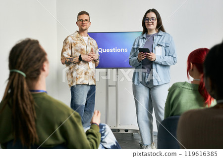 Group of people engaging in QA session during conference event with presenters standing in front holding tablet and microphone, with audience raising hands for questions 119616385