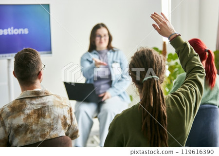 Group of participants engaging in interactive business seminar, with one individual raising hand while seated and another person presenting in background with laptop Group of participants engaging in interactive business seminar, with one individual raising hand while seated and another person presenting in background with laptop 119616390
