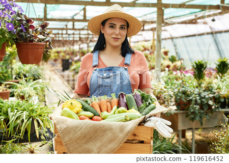 Portrait of Hispanic woman in overalls and straw hat proudly showing fresh-picked vegetables in greenhouse setting showcasing vibrant plants and flowers around her Portrait of Hispanic woman in overalls and straw hat proudly showing fresh-picked vegetables in greenhouse setting showcasing vibrant plants and flowers around her 119616452