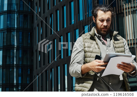 Businessman standing outside a contemporary office building analyzing paperwork held in his hands without looking at the camera Businessman standing outside a contemporary office building analyzing paperwork held in his hands without looking at the camera 119616508