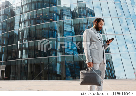 Businessman dressed in gray suit standing outside large glass office building, engrossed in smartphone while holding leather briefcase Businessman dressed in gray suit standing outside large glass office building, engrossed in smartphone while holding leather briefcase 119616549