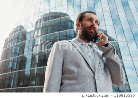 Businessman standing outside modern glass office building talking on smartphone with concentrated expression 119616550