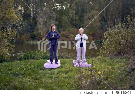 middle-aged woman teaches her old mother somatic exercises and yoga to maintain health by the river in autumn 119616841