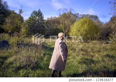 elderly woman with gray hair in light beige coat enjoying nature in autumn forest 119616877
