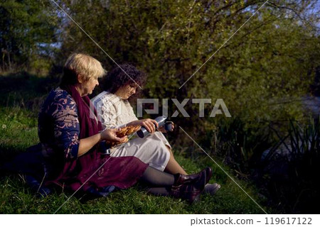 middle-aged daughter and old mother eating pastries and drinking tea from thermos cups on a picnic in the autumn forest middle-aged daughter and old mother eating pastries and drinking tea from thermos cups on a picnic in the autumn forest 119617122