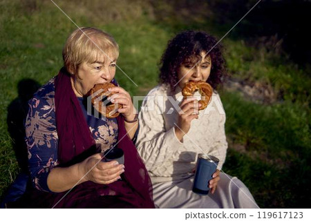 middle-aged daughter and old mother eating pastries and drinking tea from thermos cups on a picnic in the autumn forest middle-aged daughter and old mother eating pastries and drinking tea from thermos cups on a picnic in the autumn forest 119617123