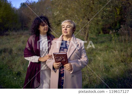 middle-aged woman and elderly mother with holy bible in hands praying for peace in autumn forest 119617127