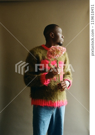 Vertical artistic portrait of young African American man holding flowers in bloom and wearing sweater with pink accent color shot on film in studio Vertical artistic portrait of young African American man holding flowers in bloom and wearing sweater with pink accent color shot on film in studio 119618451