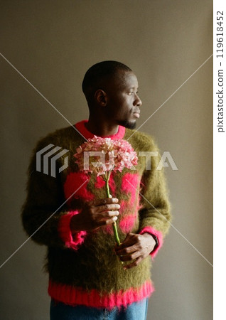Side view fashion portrait of young African American man holding pink flowers in bloom and wearing soft fuzzy sweater shot on film in studio Side view fashion portrait of young African American man holding pink flowers in bloom and wearing soft fuzzy sweater shot on film in studio 119618452